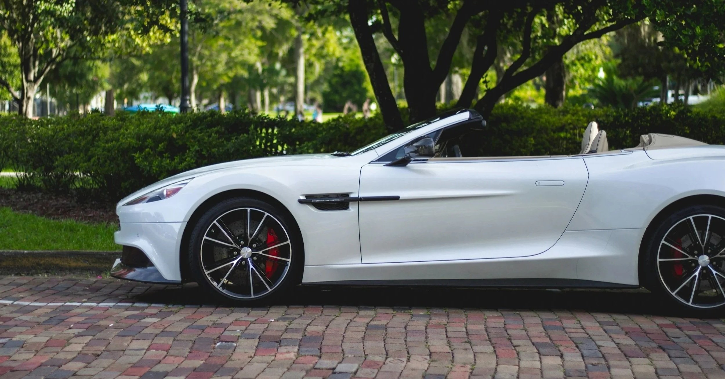 A white convertible sports car parked on a brick road in a park with green trees and bushes in the background.