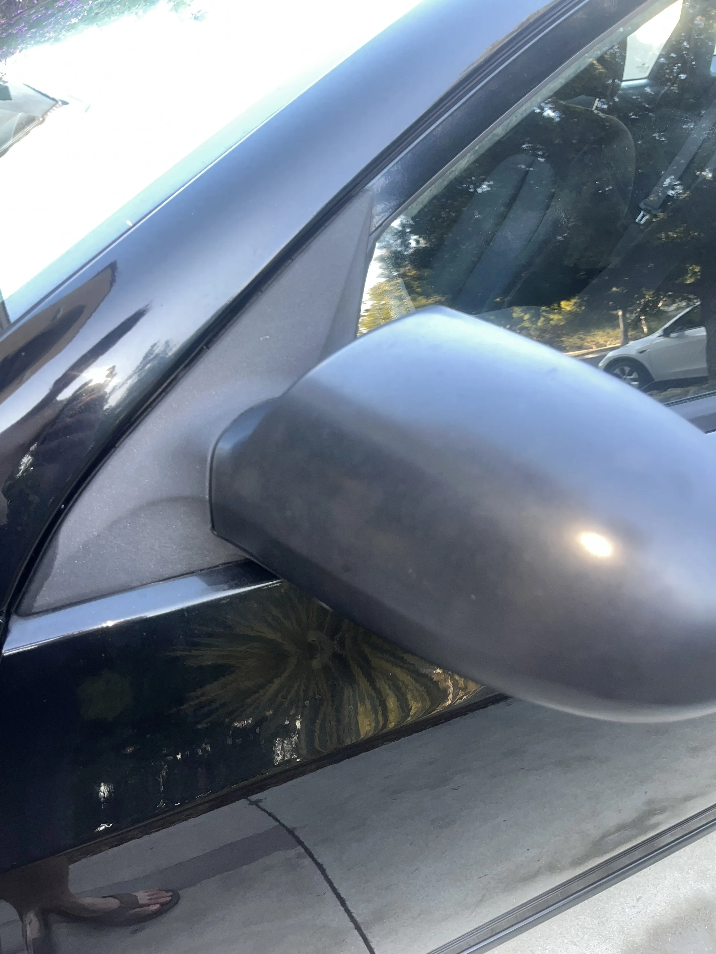Close-up of a side mirror on a black car parked outdoors, with reflections of trees and the sky on the car surface. A before photo of the effects of plastic treatment during a detail in Torrey Pines.