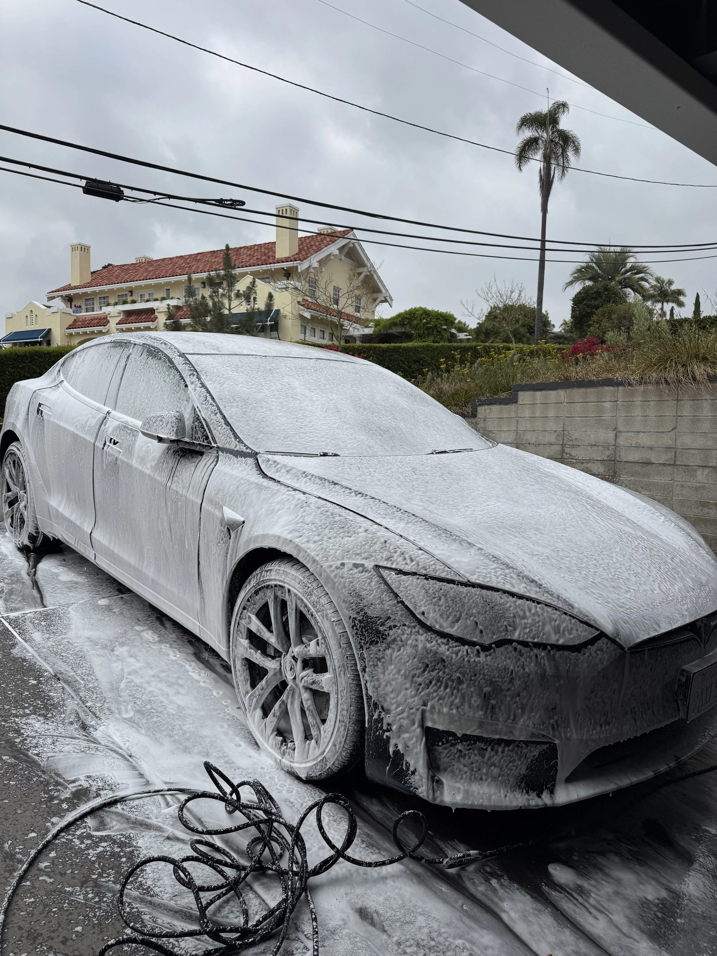 A black car covered in soap foam during a mobile car wash, with cleaning hoses and a wet concrete ground underneath, in Del Mar, California with houses, trees, and power lines in the background on a cloudy day.