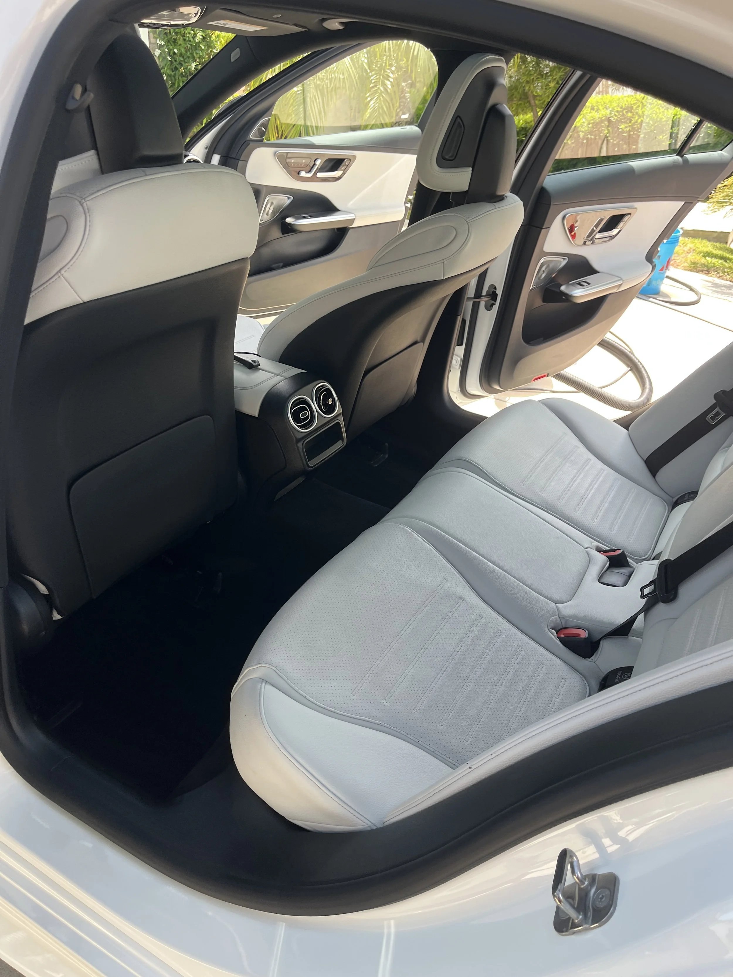 The interior of a Mercedes featuring light gray leather seats, black back of the front seats, rear air vents, and an open door showing a green garden outside. After picture of an interior detail in Torrey Pines, California.