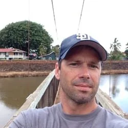 A man taking a selfie on a boat, wearing a baseball cap and a gray shirt, with a river, trees, and houses in the background.