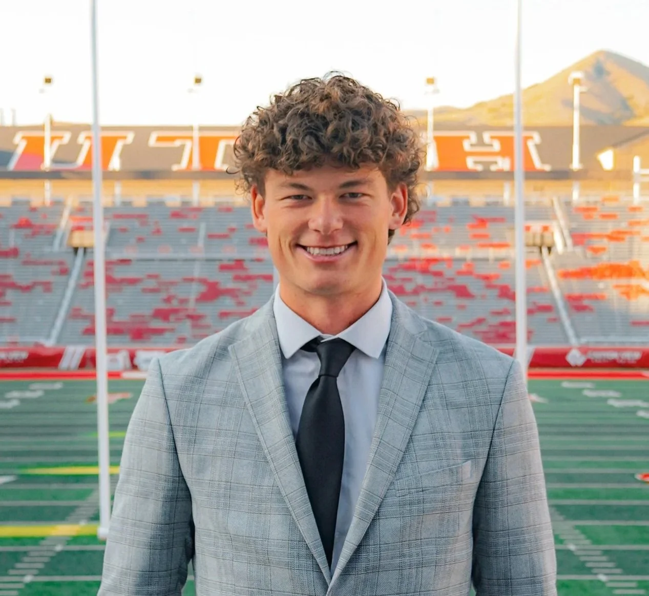 A young man in a gray suit and black tie standing on a football field in a stadium, smiling at the camera.