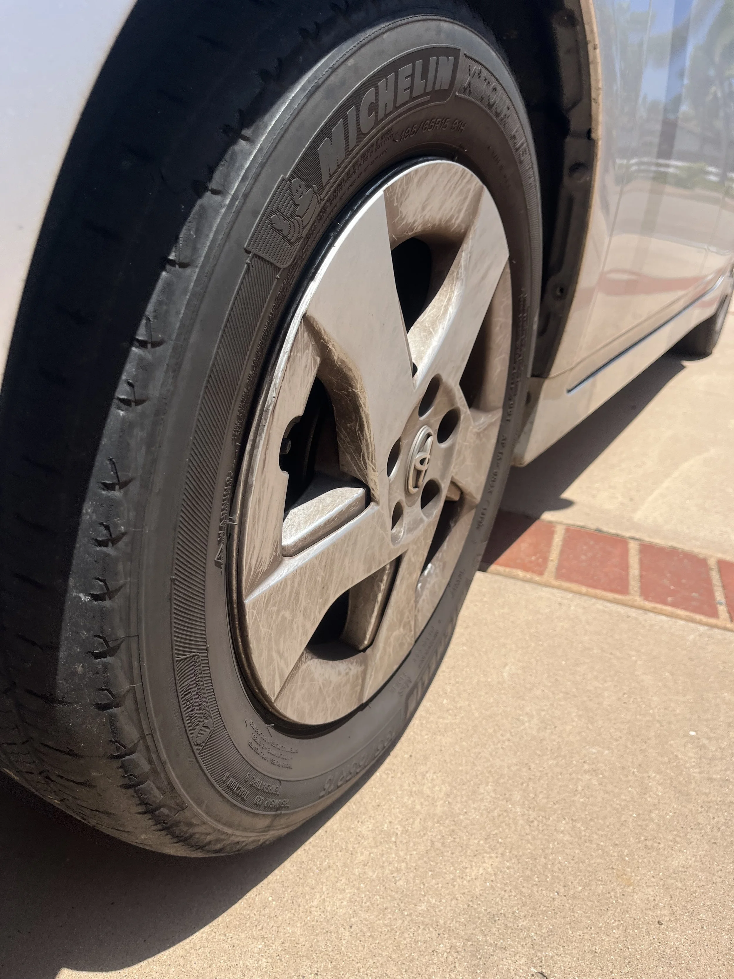 Close-up of a car tire and wheel, showing the Michelin tire brand and Chrysler wheel design, parked on a concrete driveway. Before picture of a sedan tire during a detail in poway, California.