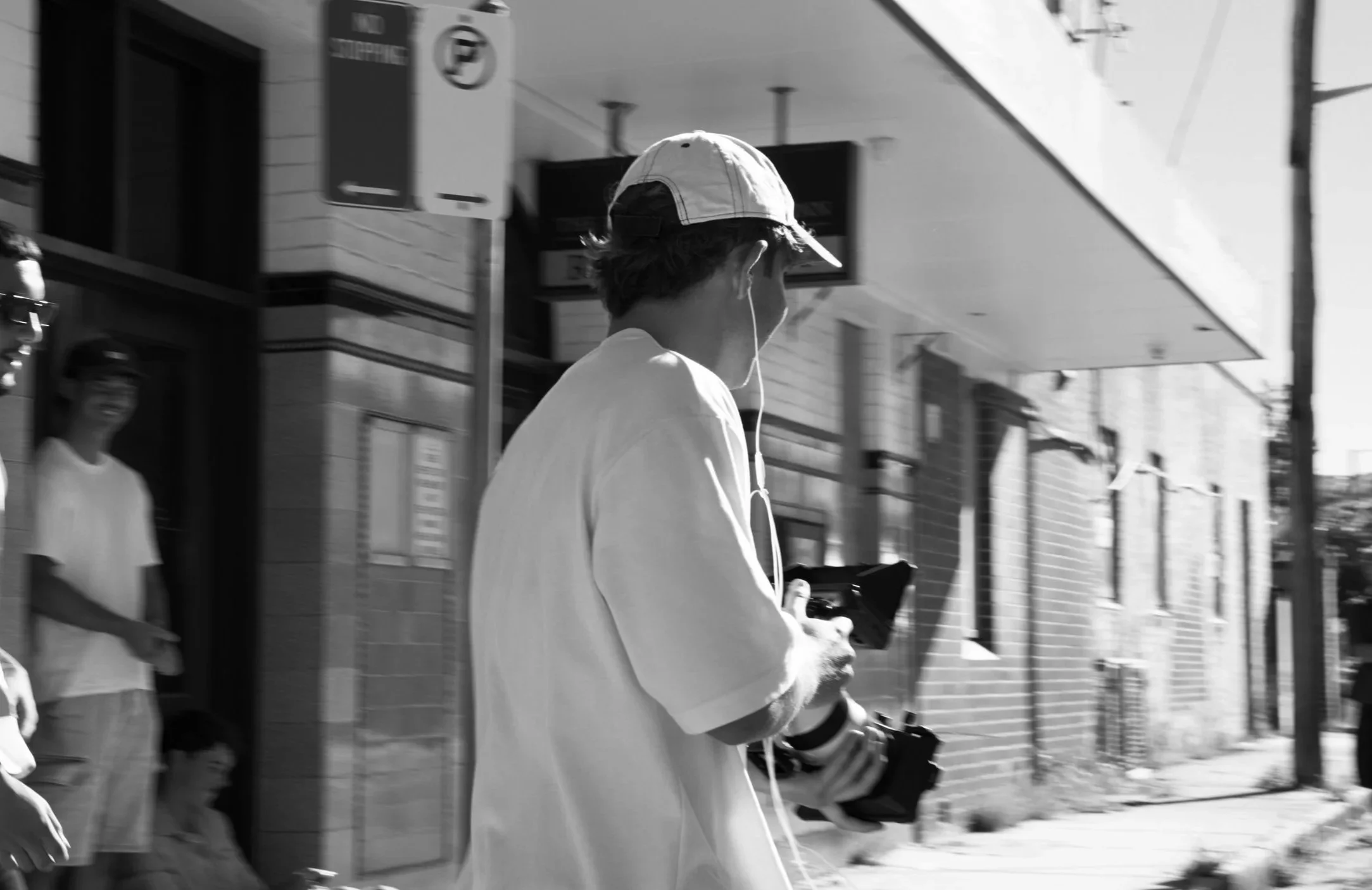 A young man wearing a baseball cap and earphones is walking on a city sidewalk while holding a camera. There are a couple of people in the background near a storefront.