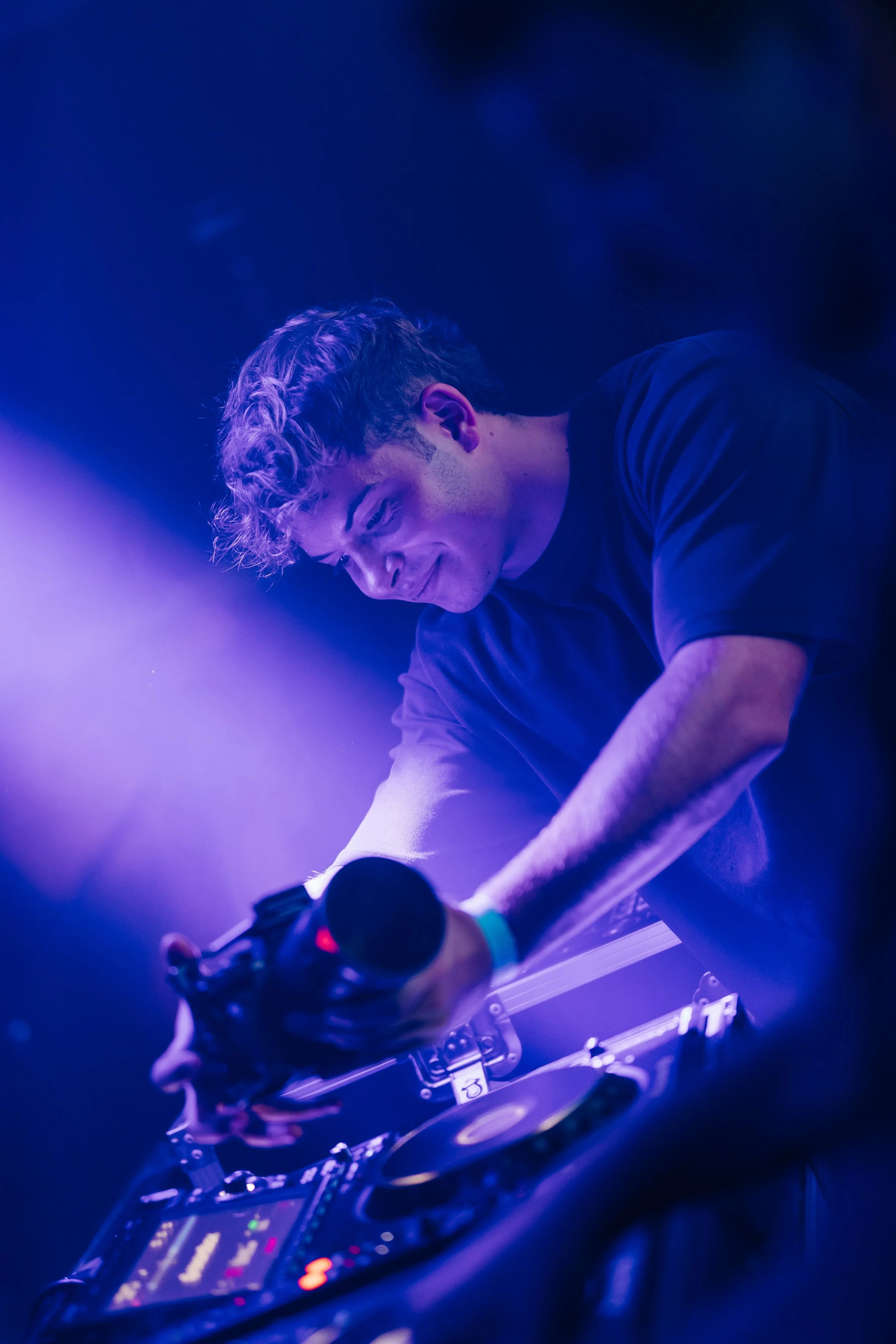 A DJ with light-colored, curly hair in a dark room illuminated by blue and purple lights, operating a turntable and DJ equipment.