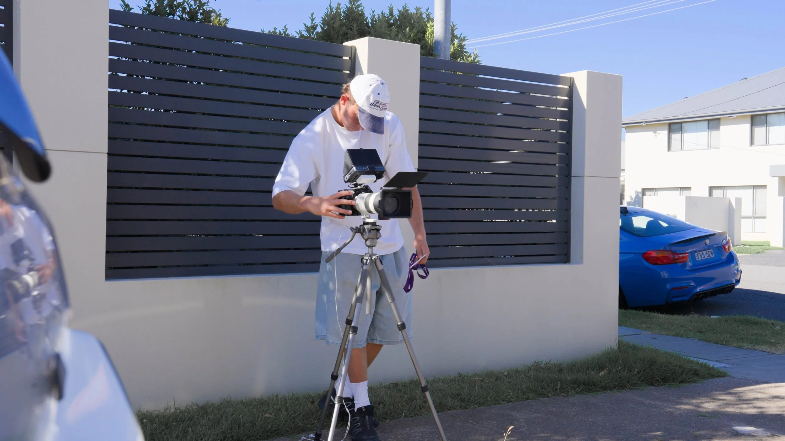A person setting up camera equipment outdoors near a modern residential area with a blue car parked beside a wall and house in the background.