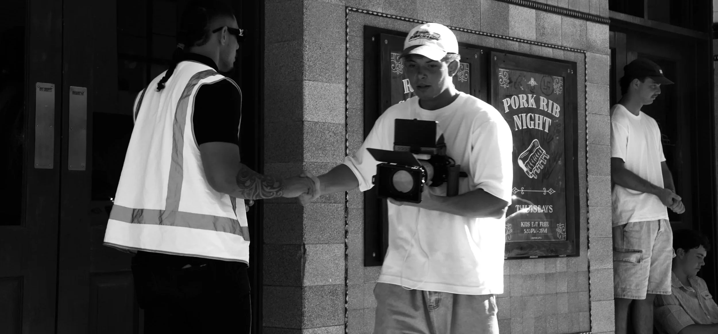 Two men are shaking hands in front of a restaurant with a sign reading 'Pork Rib Night.' One man is wearing a safety vest, and the other man is holding a professional camera. Two other men are standing in the background, one leaning against the wall and the other sitting.