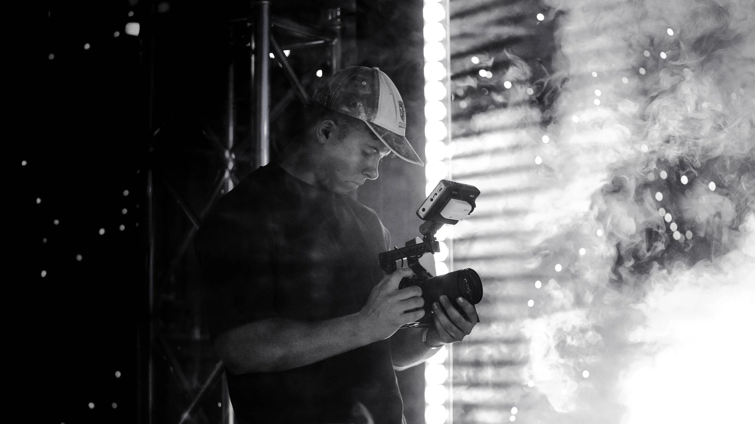 A person wearing a cap is holding a professional camera with a mounted monitor, standing in front of illuminated background lights and smoke, captured in black and white.
