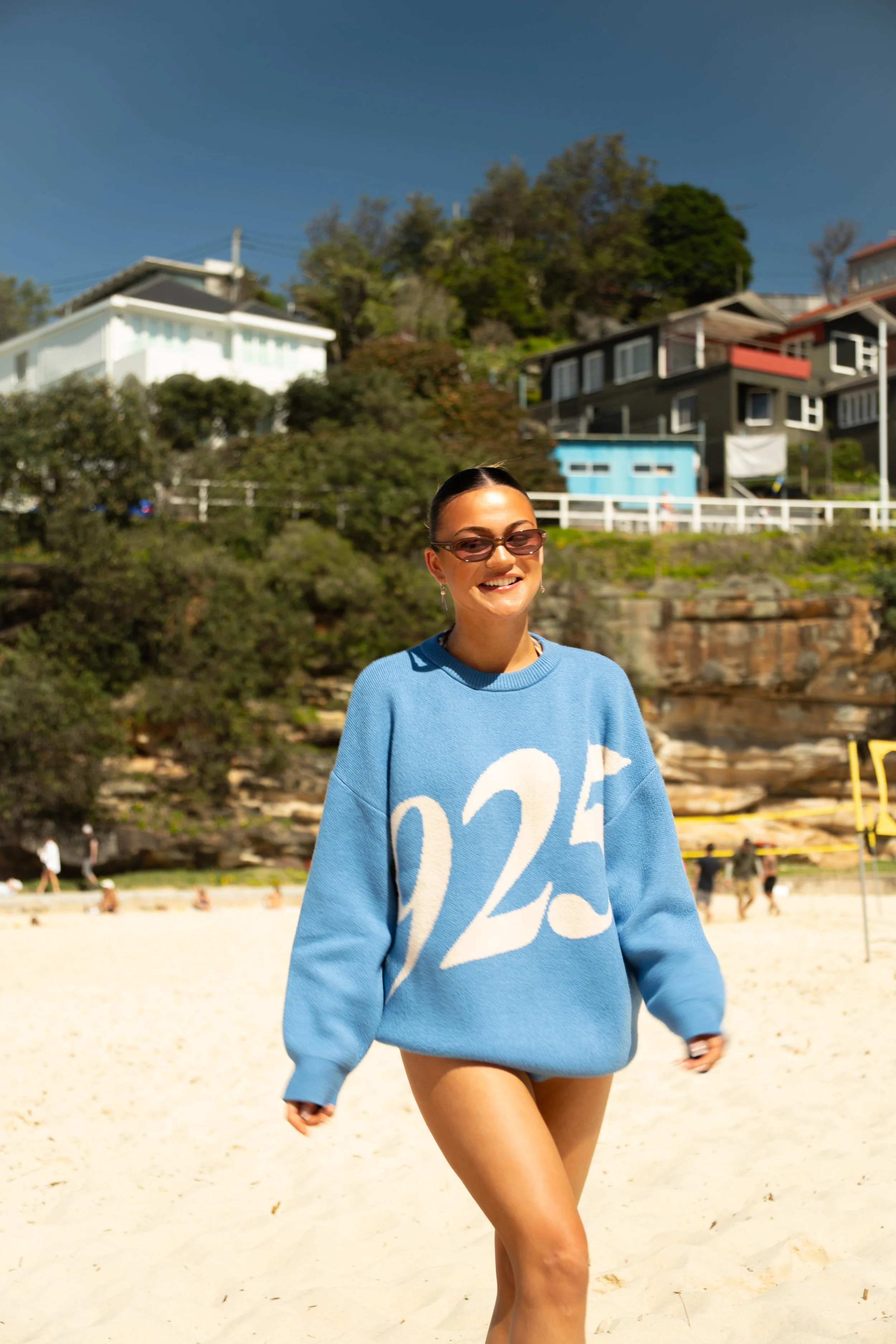 A woman smiling and walking on a sandy beach, wearing sunglasses, a blue sweatshirt with large white numbers, and no pants or shorts.