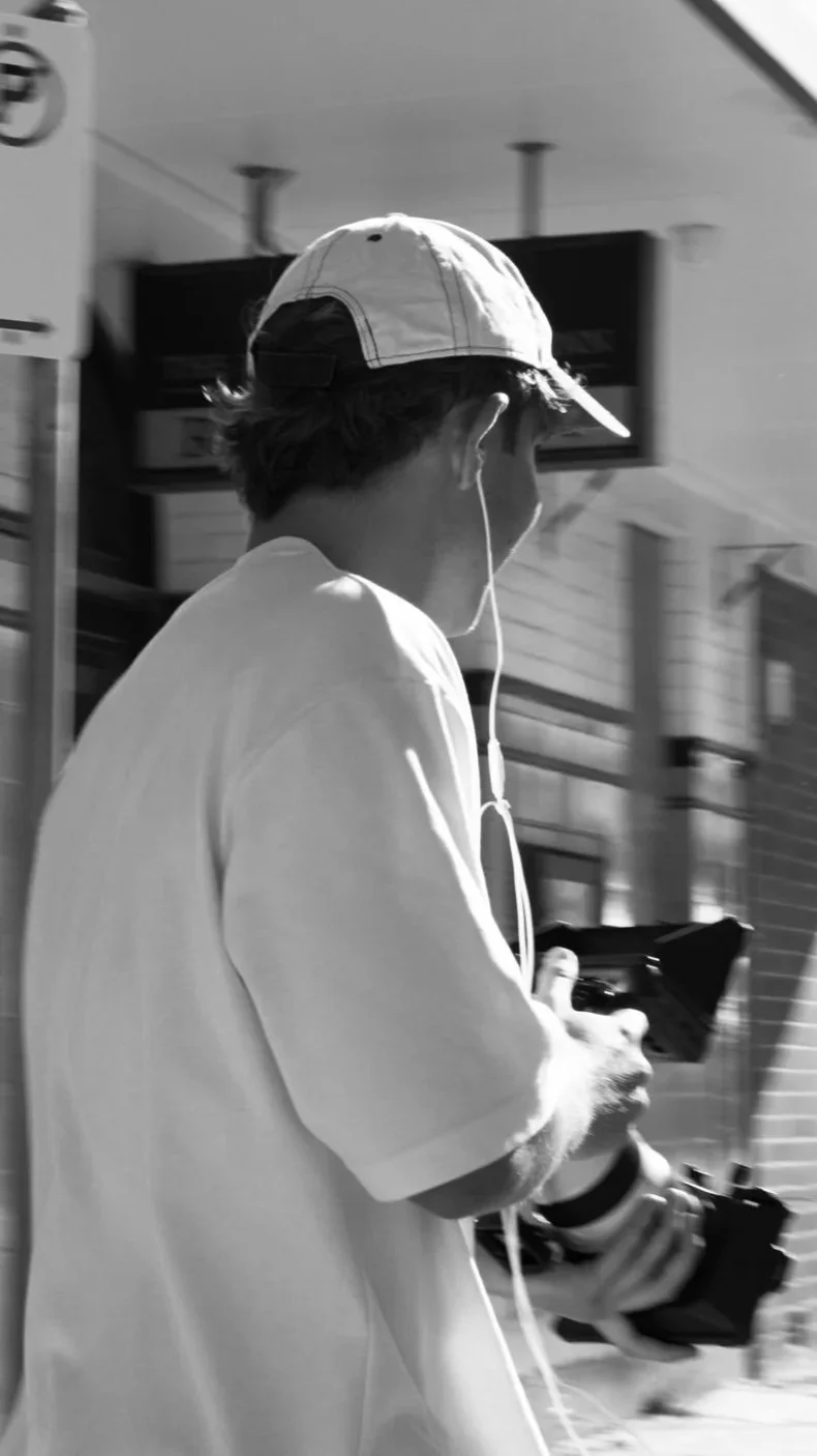 A person wearing a baseball cap and headphones is holding a camera while walking outdoors in an urban environment.