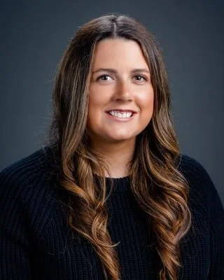 A woman with long, wavy brown hair and a black sweater smiling at the camera against a dark background.