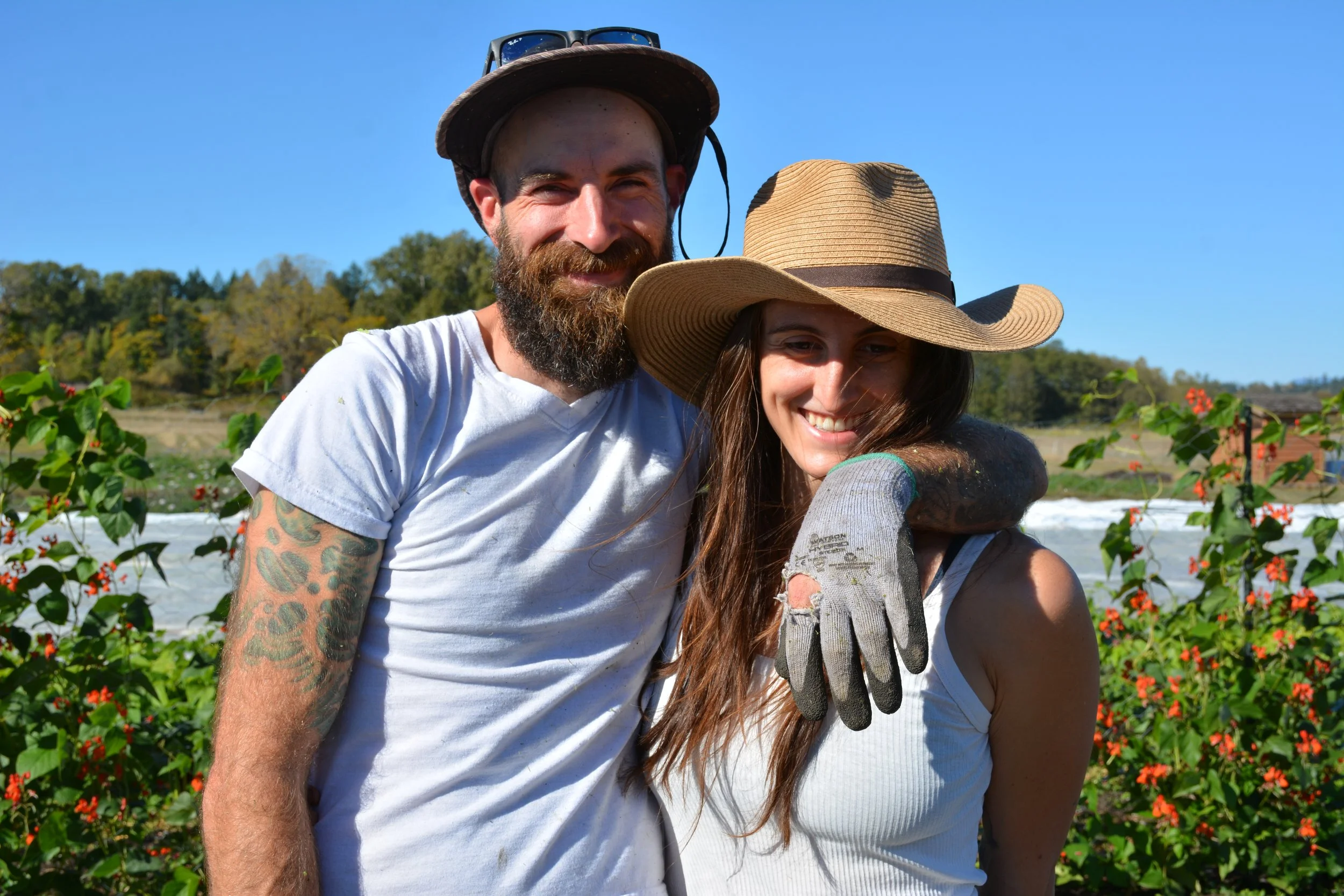 Founding members smiling in front of Testbed Farm with flowering plants, wearing hats and outdoor clothing, enjoying sunny weather.