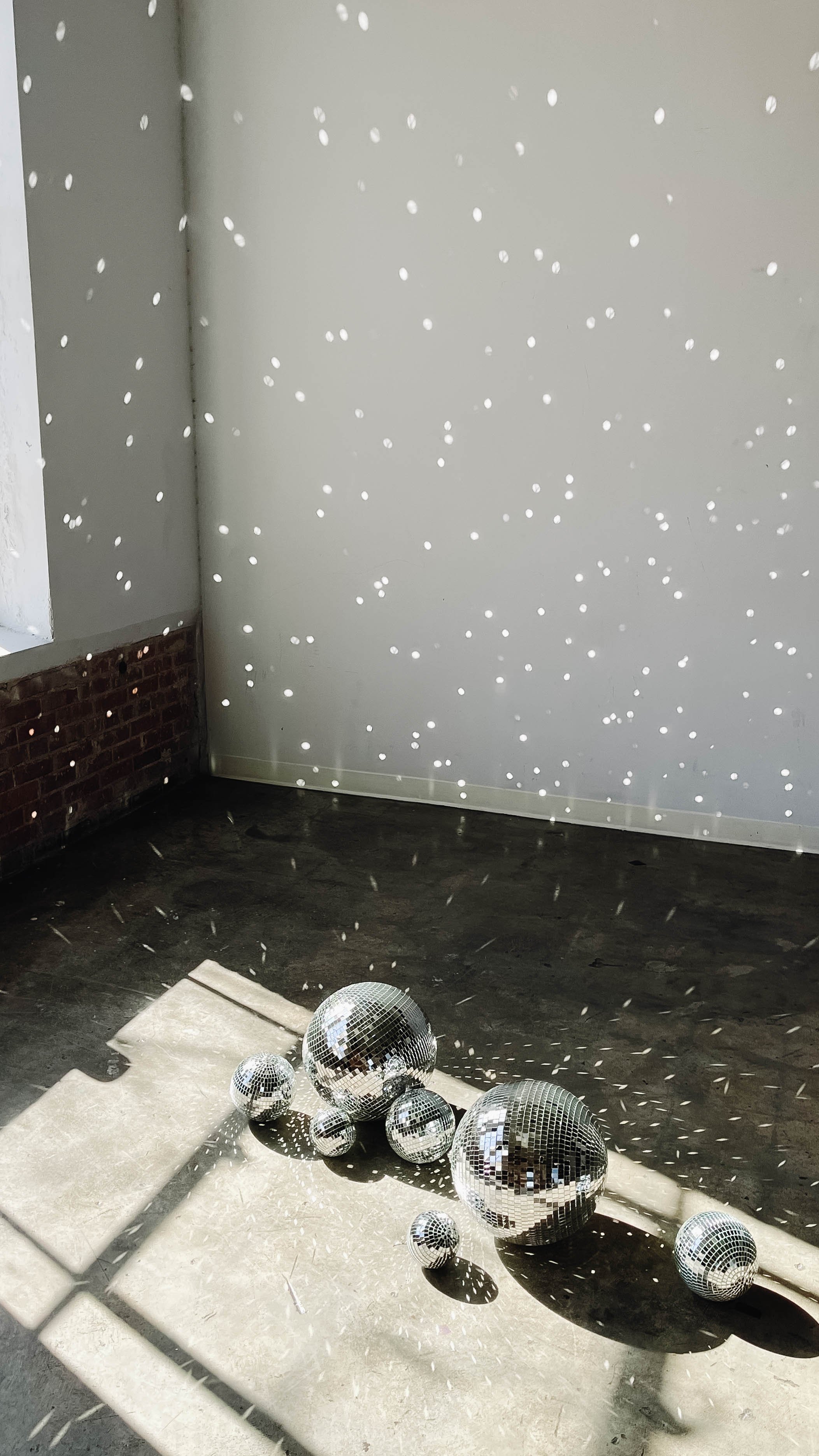 Multiple disco balls of different sizes reflecting and casting light, creating a pattern of dots on the wall and floor of a room with a brick and plain wall.
