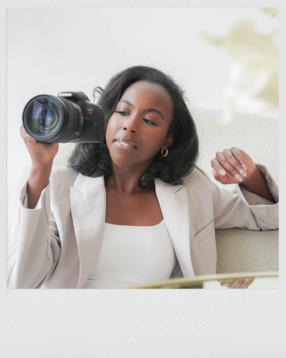 A woman with dark hair and gold earrings sitting on a light-colored sofa, holding a Canon DSLR camera and looking at the camera's screen.
