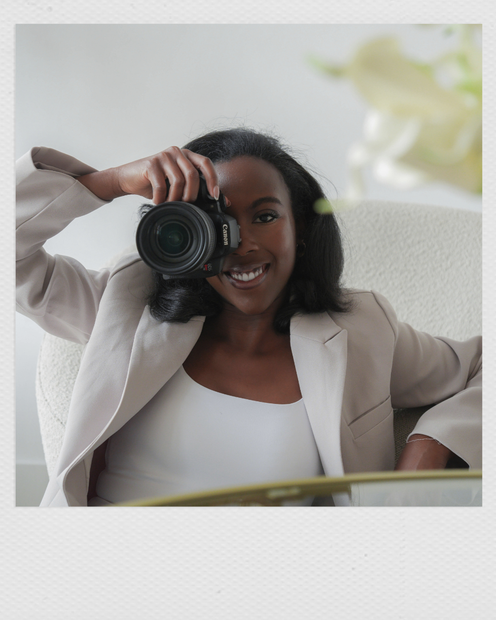 A woman smiling and looking through the viewfinder of a camera, seated on a white chair, with blurred white flowers in the background.