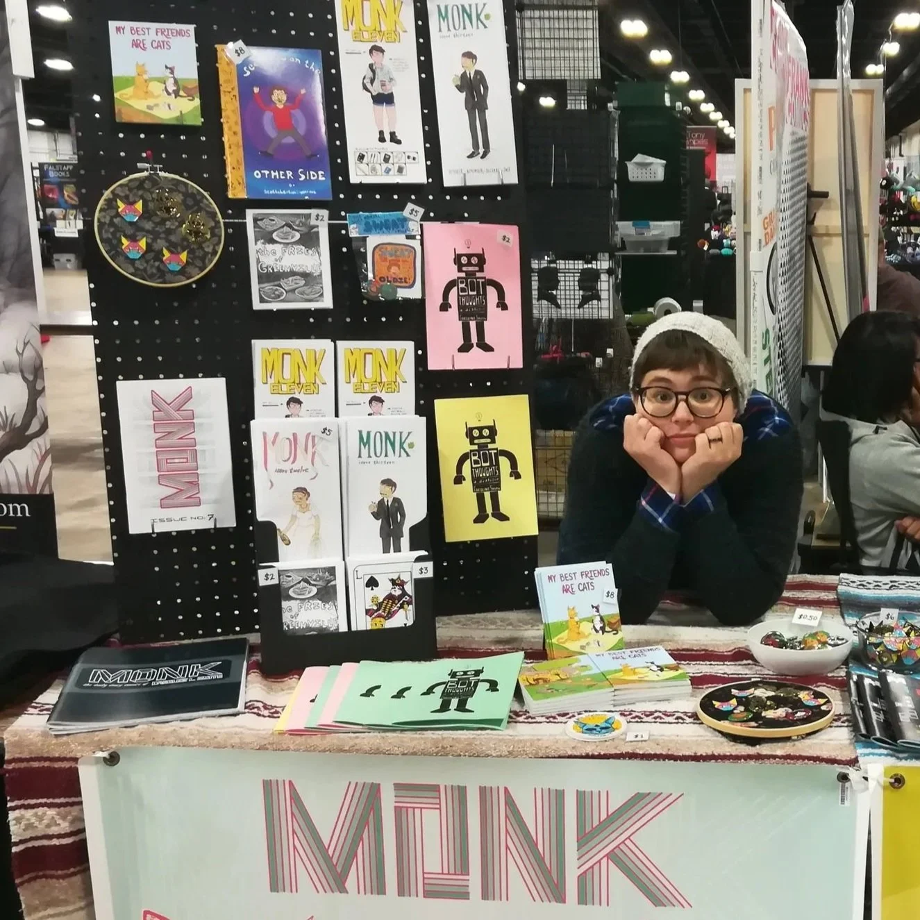 A woman with glasses and a white knit hat sitting at a booth displaying various comic and art prints, stickers, and small items in a convention or market setting.