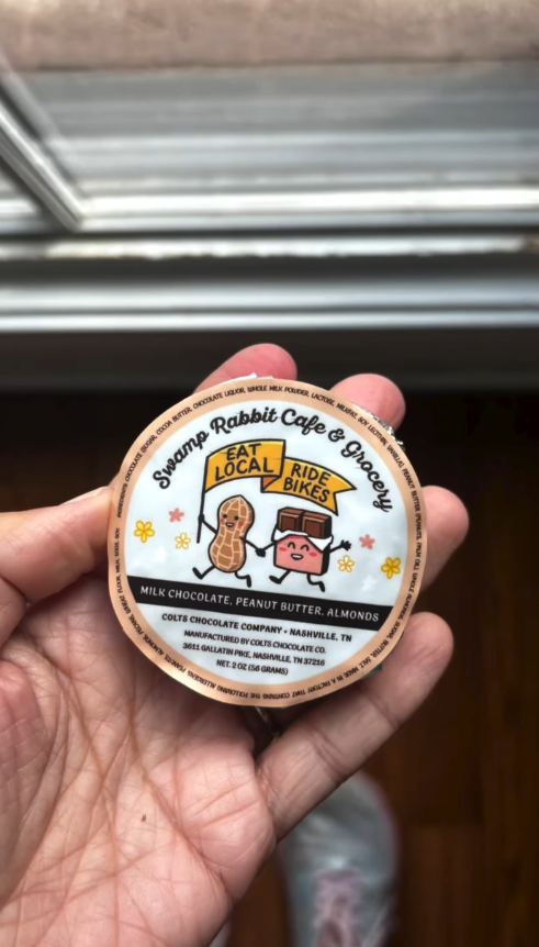 Hand holding a round chocolate wrapper with cartoon characters of a peanut and a block of chocolate, featuring the text 'Swamp Rabbit Cafe & Grocery', 'Eat Local', and 'Ride Bikes'.