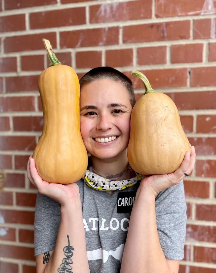 A smiling woman standing in front of a brick wall holding two large butternut squashes, one on each shoulder.