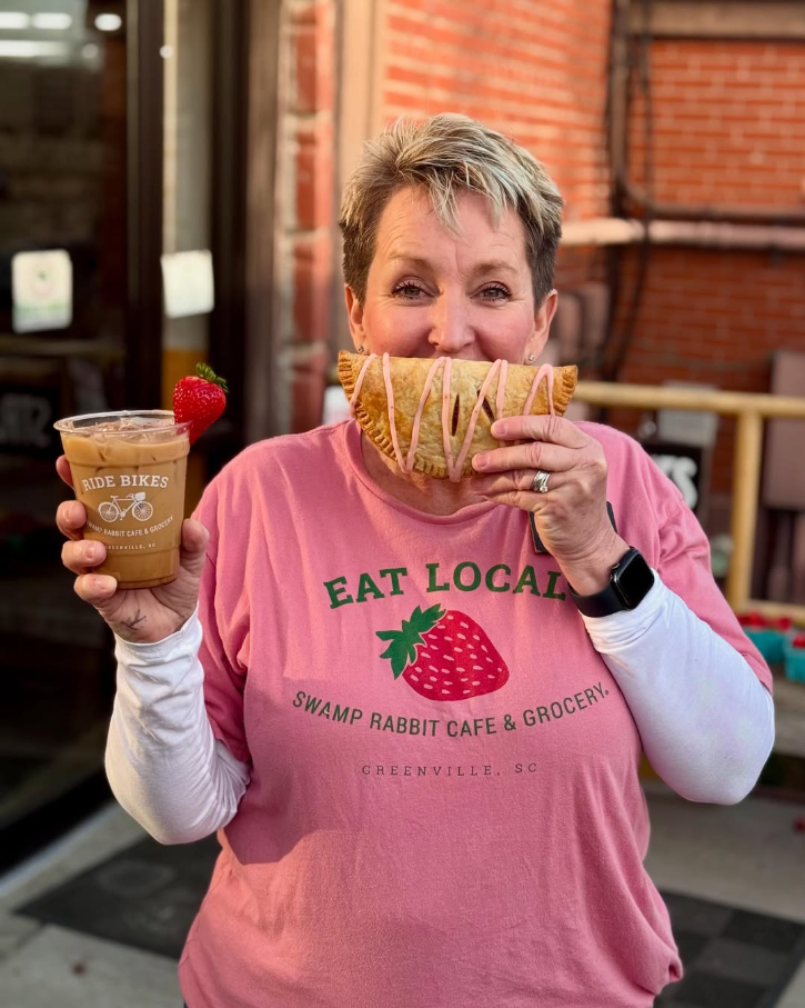 A woman holding a cup of iced coffee and a large breakfast pastry. She is wearing a pink shirt with a strawberry graphic that reads 'Eat Local' and 'Swamp Rabbit Cafe & Grocery' in Greenville, SC.