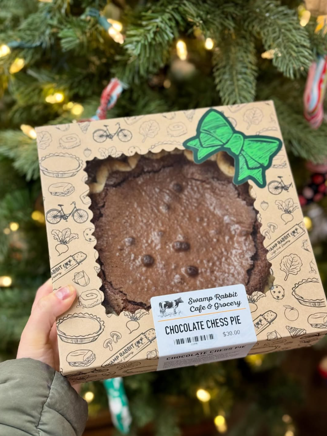 A chocolate chess pie inside a decorative box with a green bow, held in front of a Christmas tree with lights and ornaments.