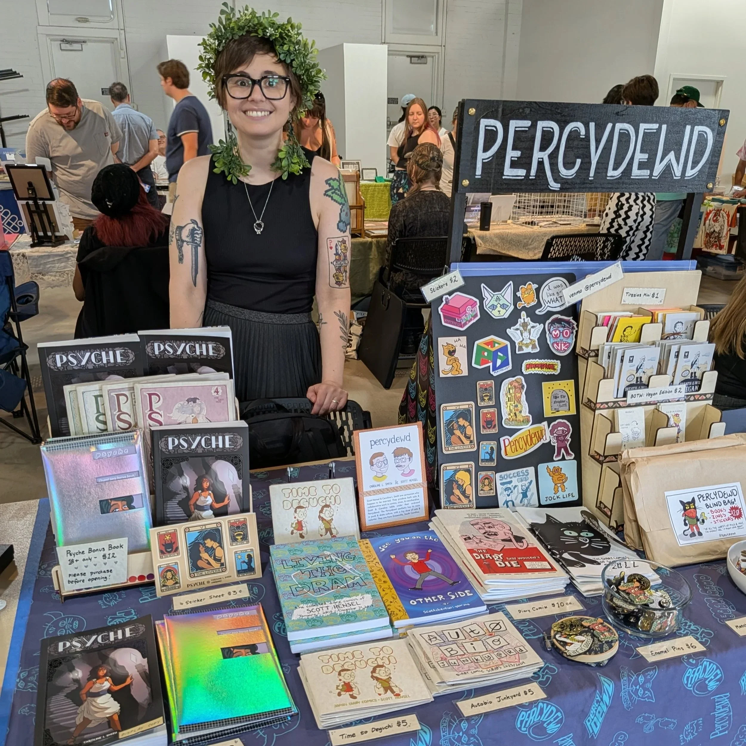 A woman with short brown hair, glasses, and tattoos on her arms stands behind a table at a booth selling comics, stickers, and pins, all related to the podcast 'PERCYWED'. She wears a black sleeveless top and a leafy crown. The booth has various items including books, stickers, and buttons displayed on the table and a sign with 'PERCYWED' on it.