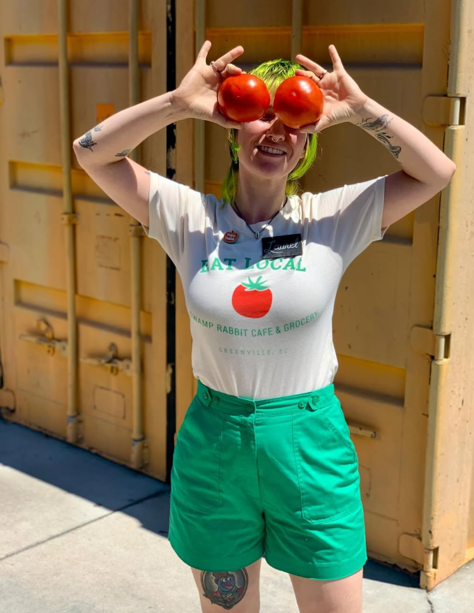 A woman with bright green hair and tattoos holding two red tomatoes over her eyes, smiling. She wears a white T-shirt with a tomato graphic and green shorts, standing in front of a yellow industrial door.