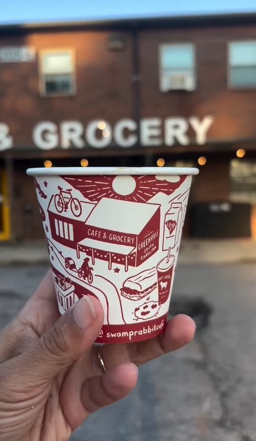A paper cup with illustrations of a cafe and grocery store, held by a person in front of a grocery store with a blurry sign reading 'GROCERY' in the background.