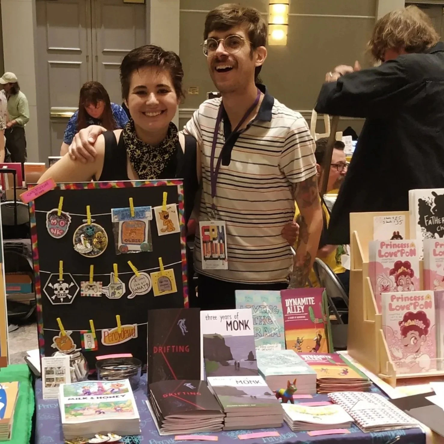 Two people standing behind a table at a book fair, smiling at the camera. The woman on the left has short, dark hair and wears a black sleeveless top with a patterned scarf. The man on the right has glasses, brown hair, and a striped shirt. The table in front displays various books, including children's books and graphic novels, and a black board with hanging pins and charms.