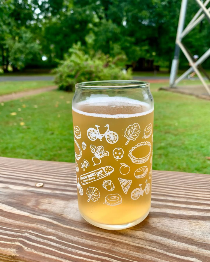 A glass of pale beer with a frothy head, decorated with white illustrations of bikes, leaves, and food items, placed on a wooden table outdoors with green trees and a cloudy sky in the background.