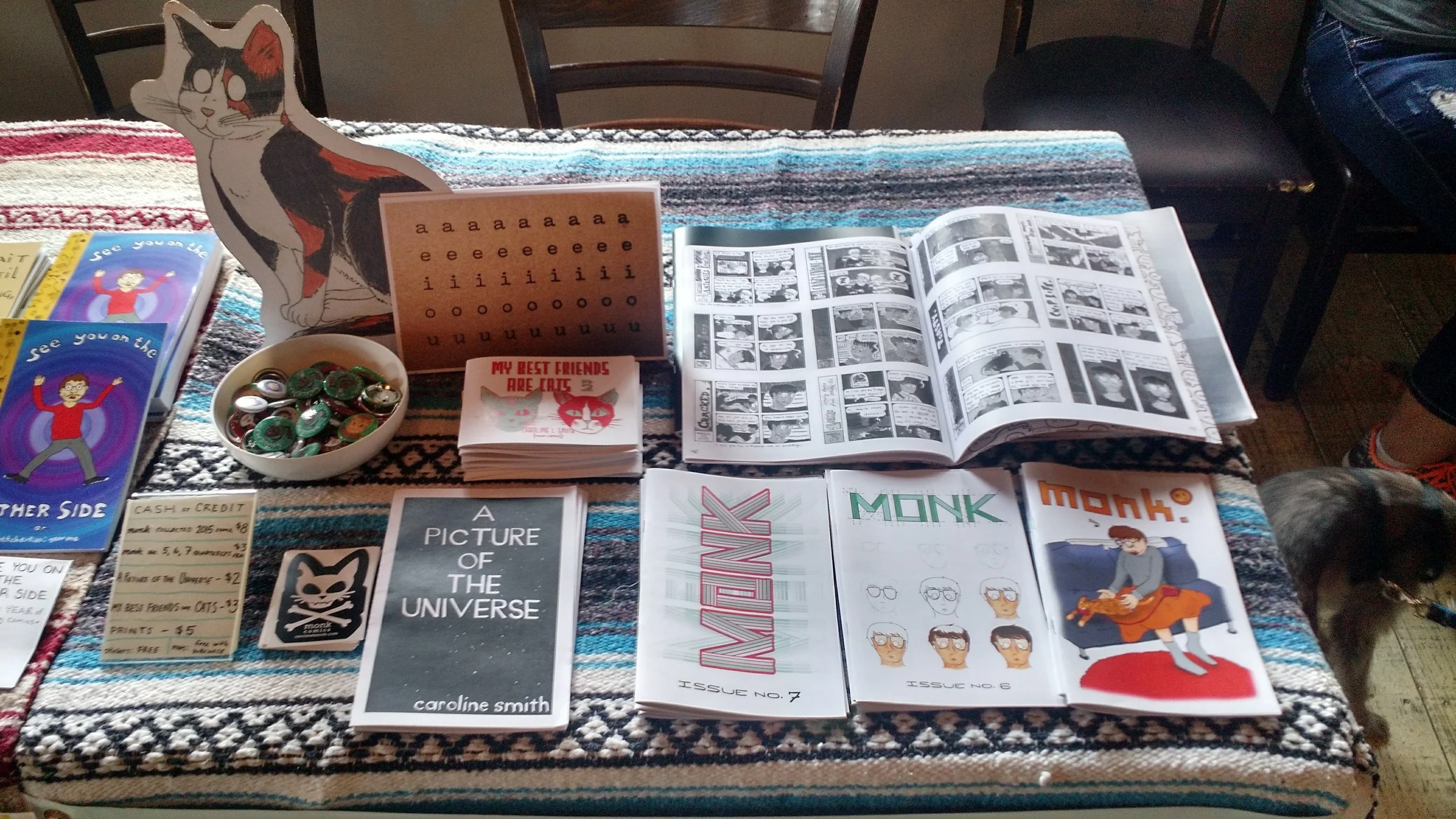 A table displaying various comics, zines, buttons, and stickers, including a clown fish cutout, a bowl of buttons, and pamphlets with alternate comic covers, on a patterned tablecloth.