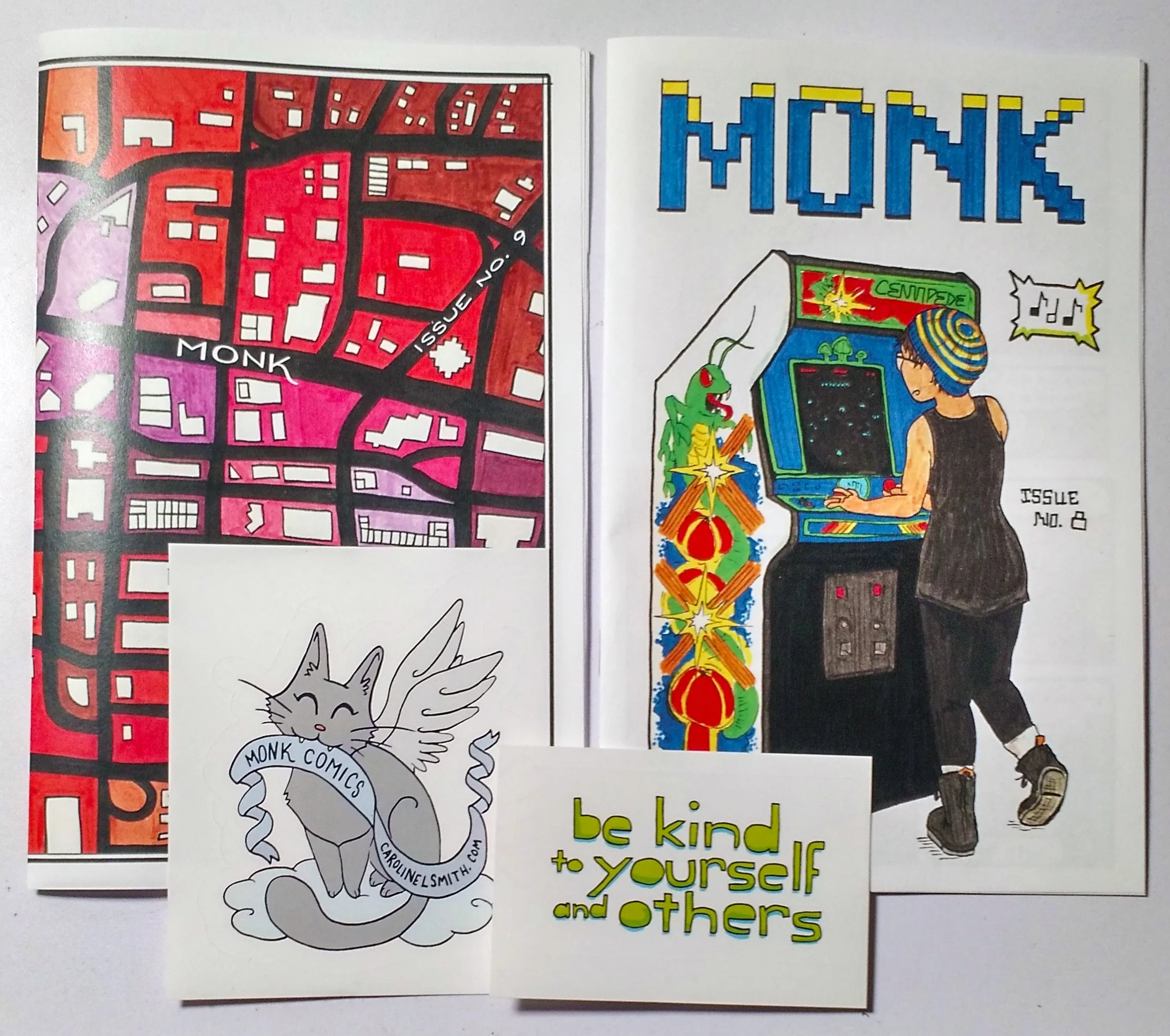 Close-up of a comic book cover with a maze map, a sticker of a winged cat with a banner reading 'Monk Comics' and a website, a coloring page of a girl playing arcade games titled 'Monk,' and a small card with the message 'be kind to yourself and others.'