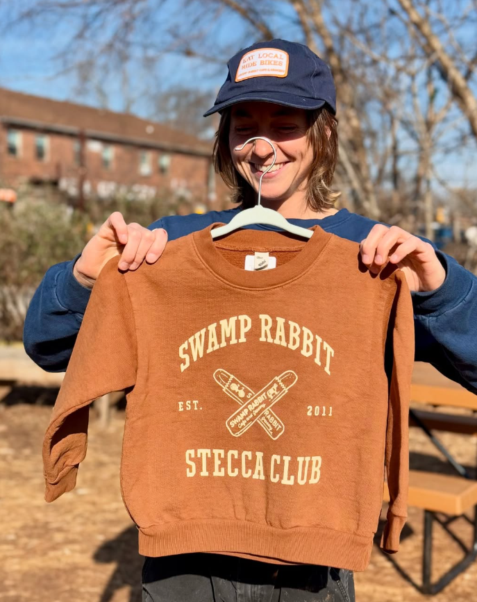 Person outdoors holding a brown sweatshirt with 'Swamp Rabbit SteeClub' printed on it, smiling, wearing a blue cap with an orange patch, clear sky and trees in the background.