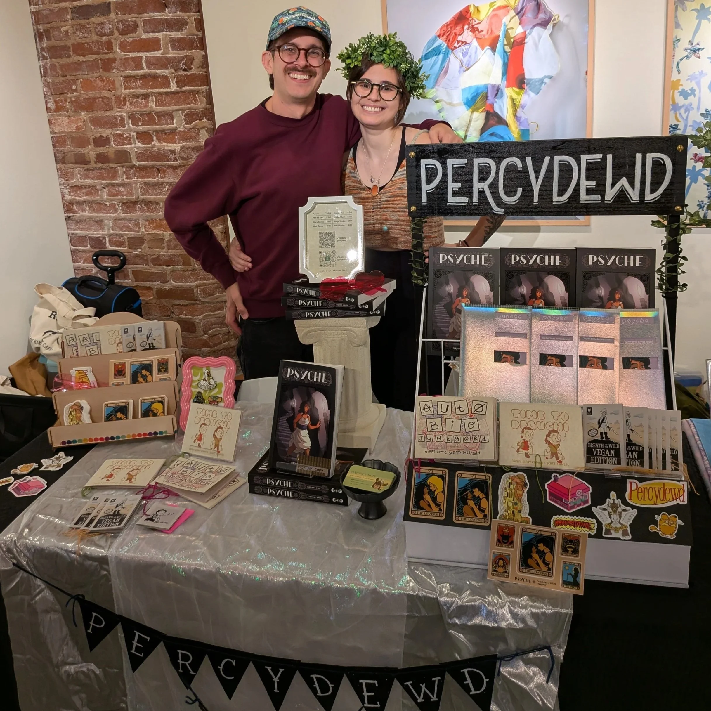 Two people standing behind a table at a booth labeled 'PERCYDEWD' at a comic or art fair, with various merchandise including art prints, cards, and comic books displayed on the table.
