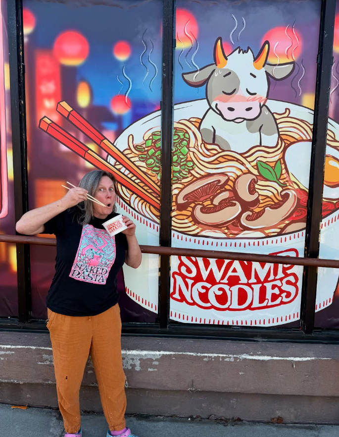 A woman with gray hair, wearing a black t-shirt with colorful art and orange pants, is holding a box of instant ramen and using chopsticks to eat in front of a large mural of steaming ramen noodles and a cartoon cow with a happy expression.