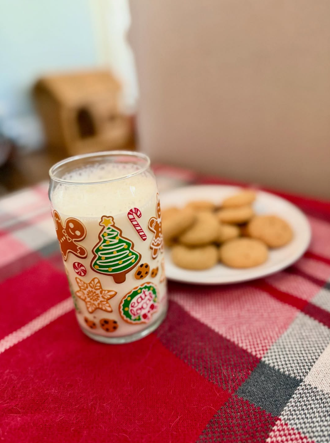 A glass with Christmas decorations containing a beverage, and a plate of cookies on a red and black checkered tablecloth.