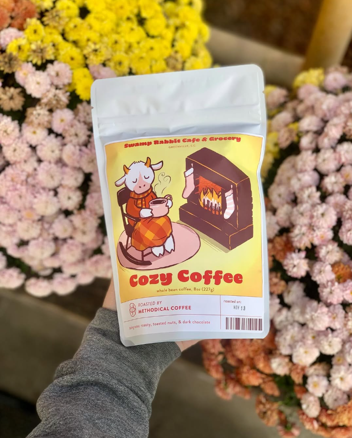 Bag of Cozzy Coffee in front of pink and yellow flowers.