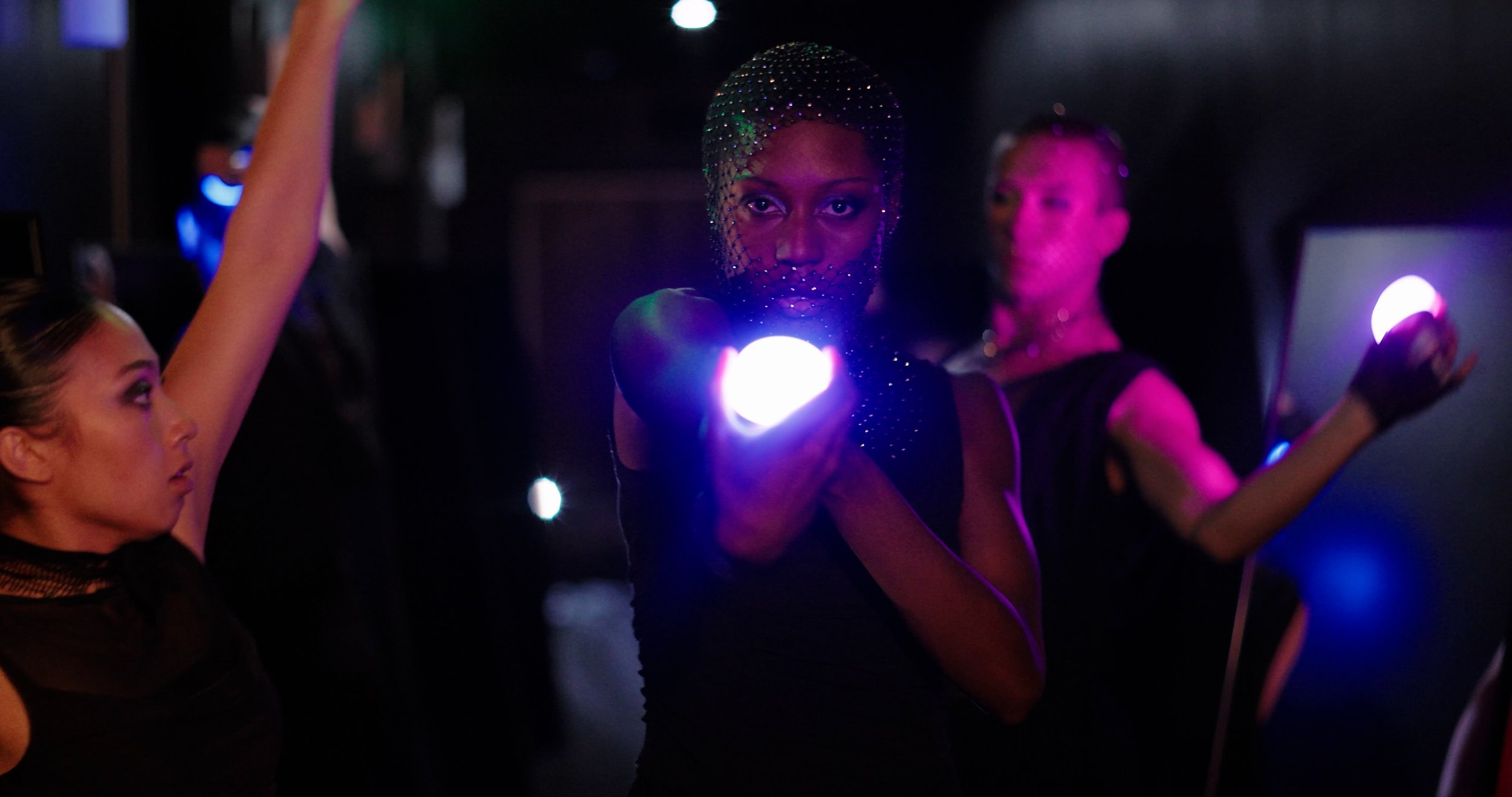 Three dancers in black outfits with illuminated gloves and accessories, dancing in a dark room with colorful lights.
