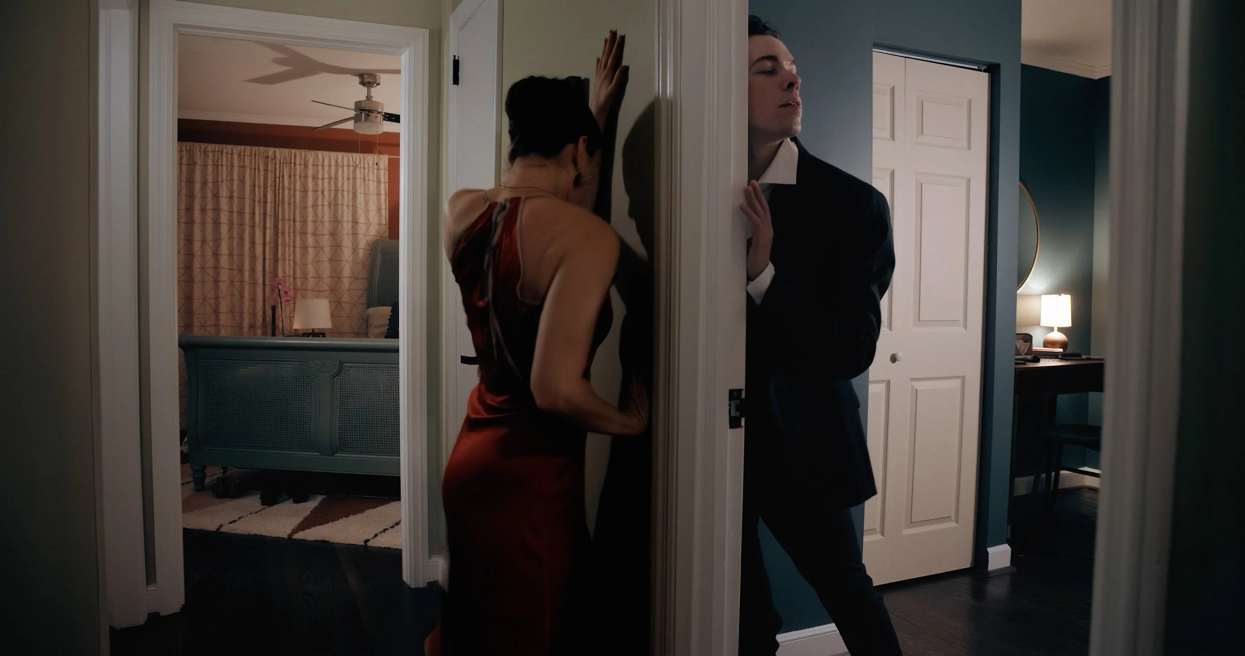 A woman in a red dress and a man in a black suit are standing on either side of a narrow doorway, appearing to be in emotional turmoil.