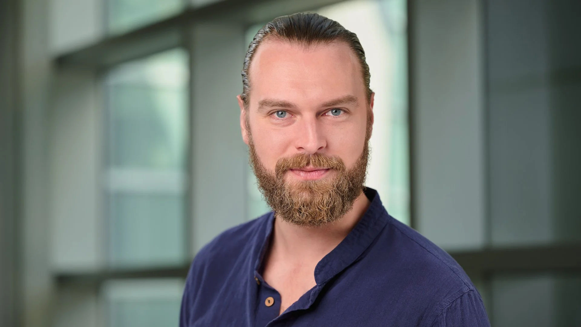Adam Witmer, a man with a beard, blue eyes, and brown hair slicked back, wearing a navy blue shirt, standing in front of a modern building with large windows.