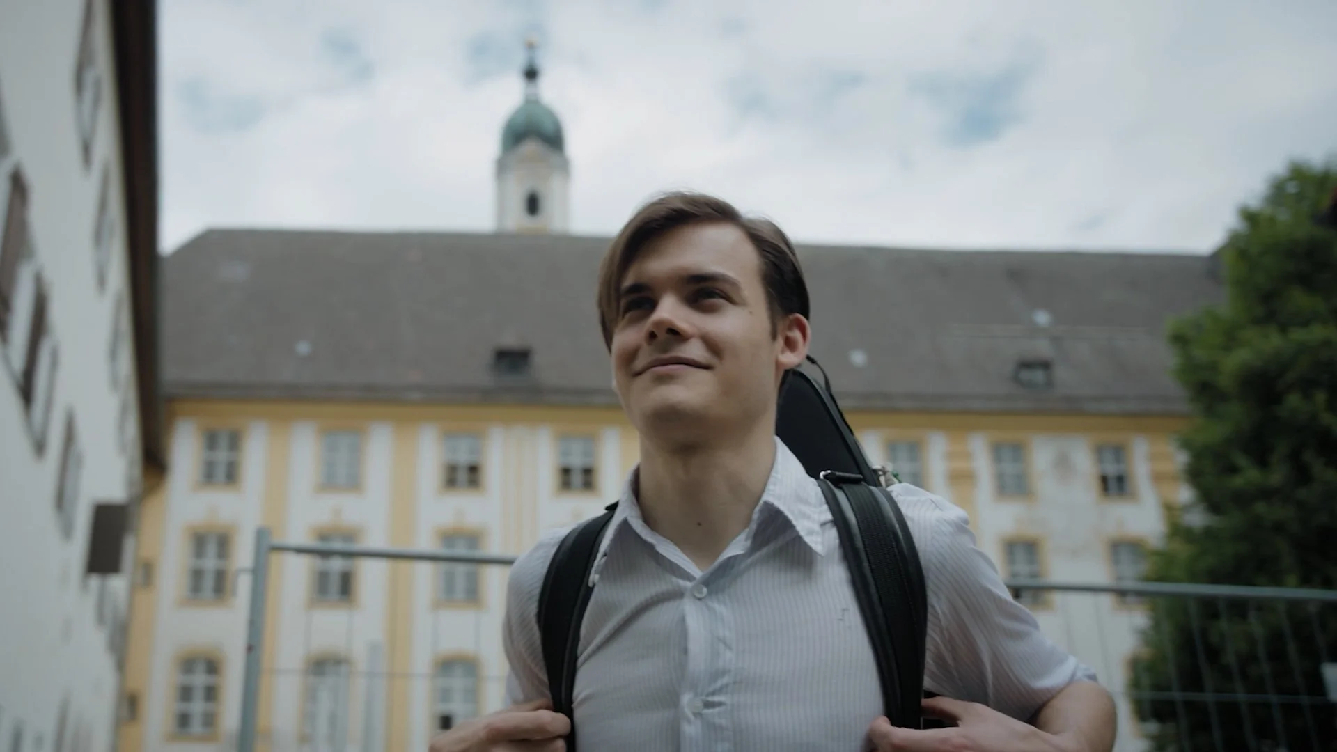 A young man with brown hair and a white shirt carrying a backpack, smiling outdoors with a historic building and church in Germany.
