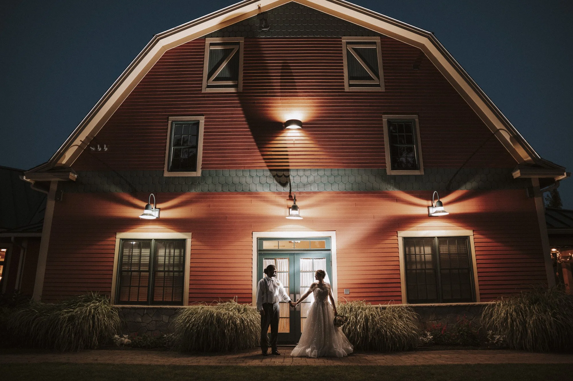 A bride and groom holding hands in front of a large red house with exterior lighting at night.