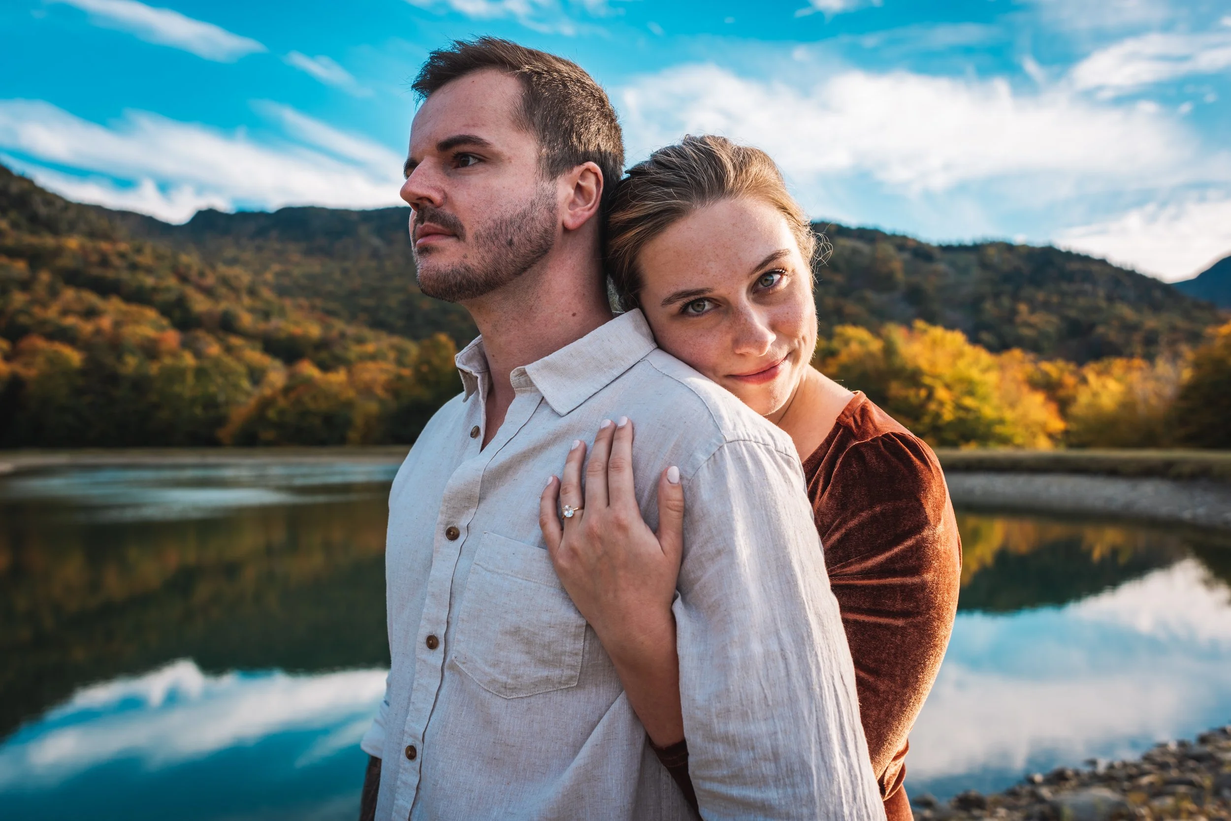 A couple embracing near a lake with mountains and trees in Stowe Vermont the background on a clear day.