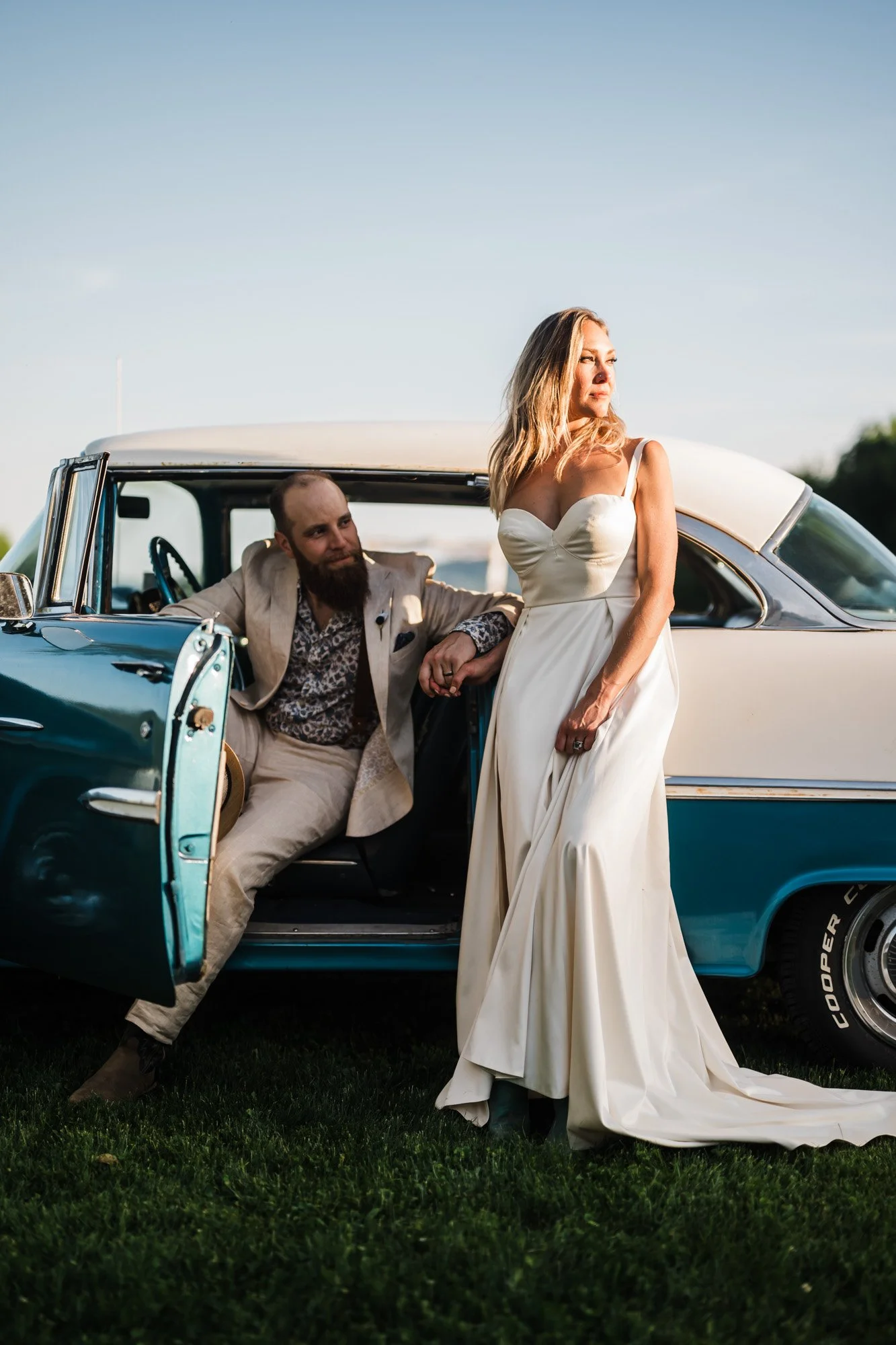 A woman in a white wedding dress standing next to a vintage blue and white car with a man sitting inside, holding her hand. The scene appears to be outdoors during the daytime.