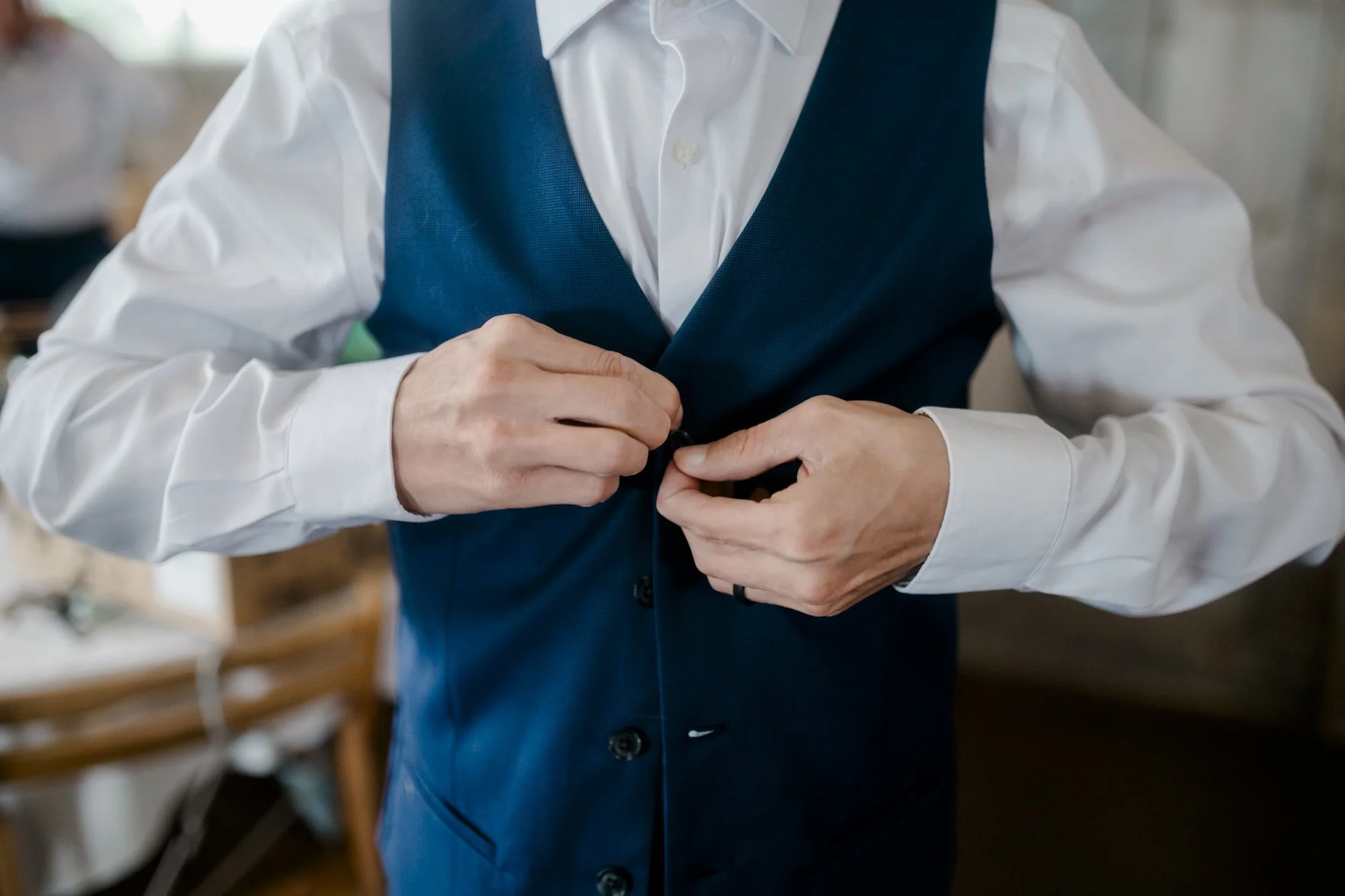 Person in white dress shirt and navy vest buttoning the vest.