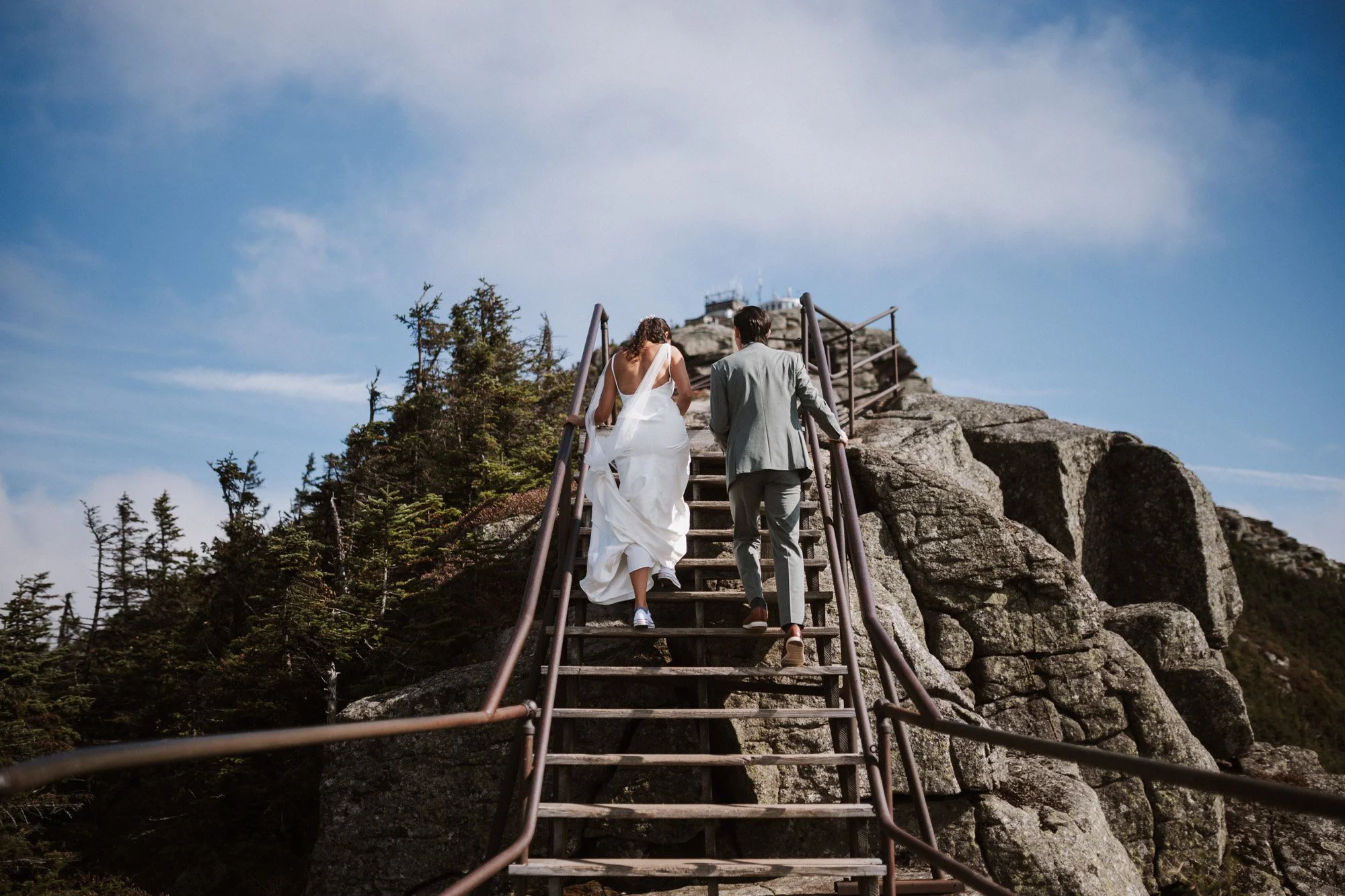 A bride and groom walking up a mountain staircase, holding hands, with a blue sky and rugged rocks in the background.