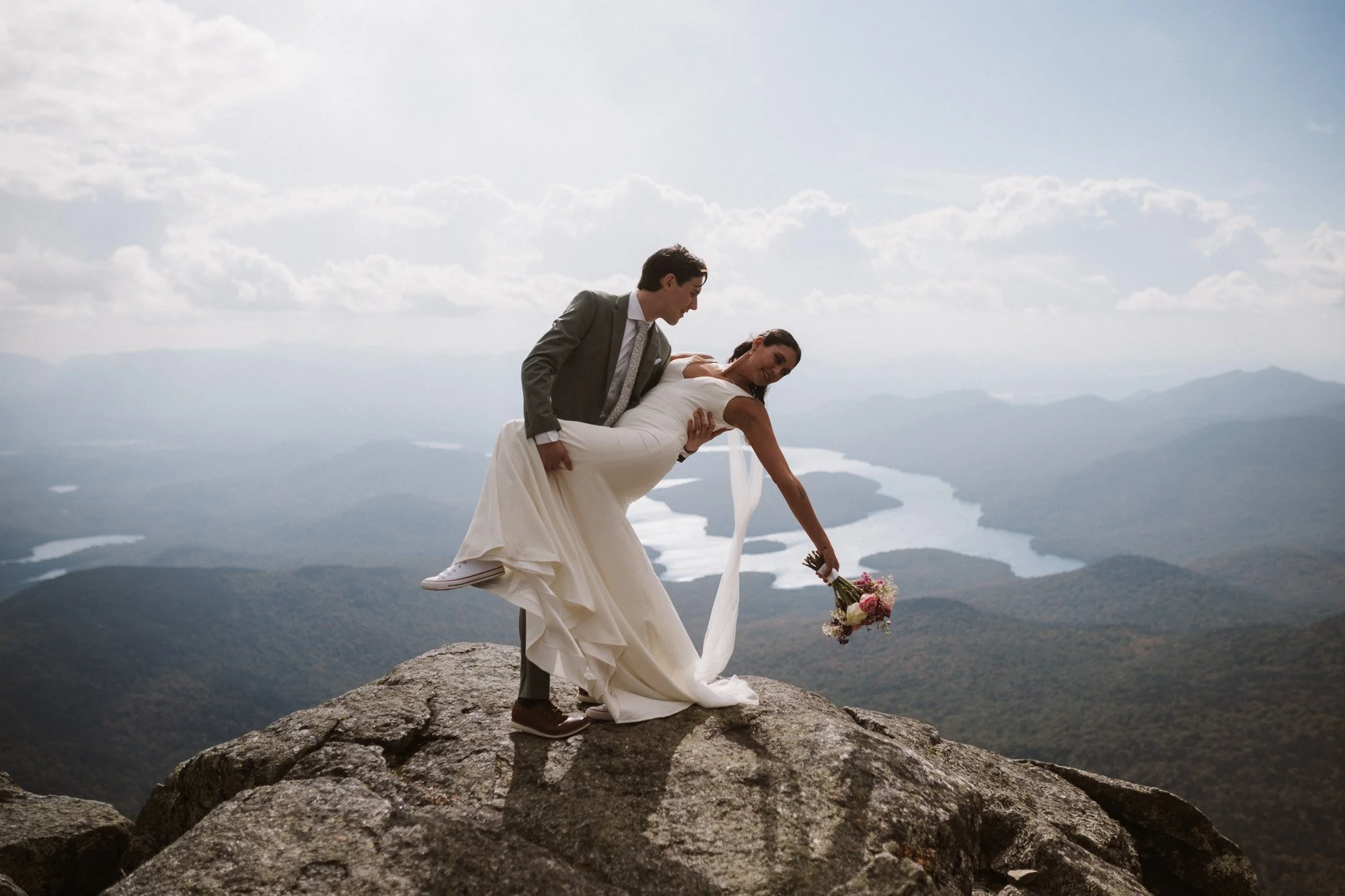 A bride and groom on a mountain peak with a scenic landscape of lakes and mountains in the background.