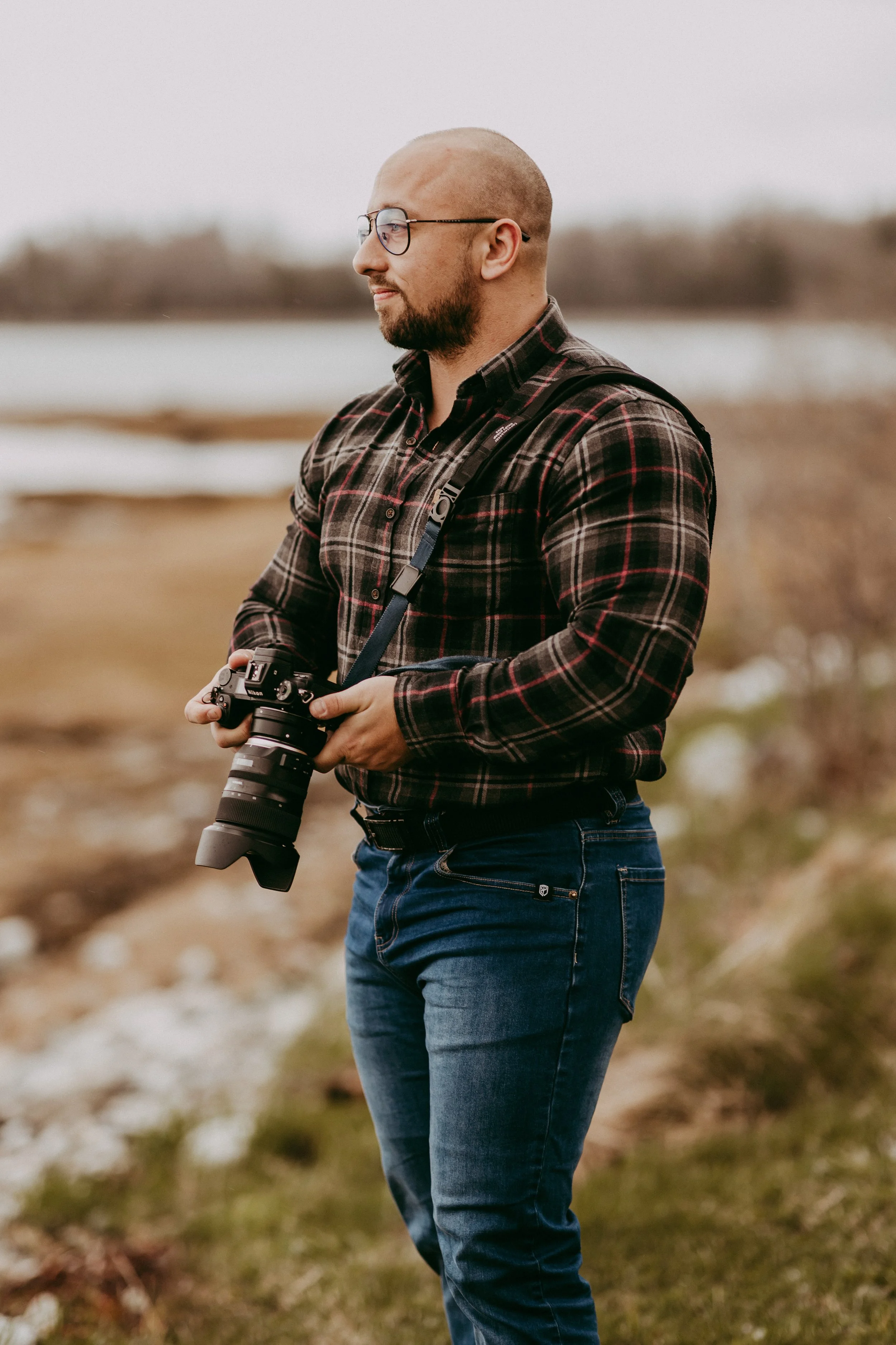 A man with a camera, wearing glasses and a plaid shirt, standing outdoors near water and grass.