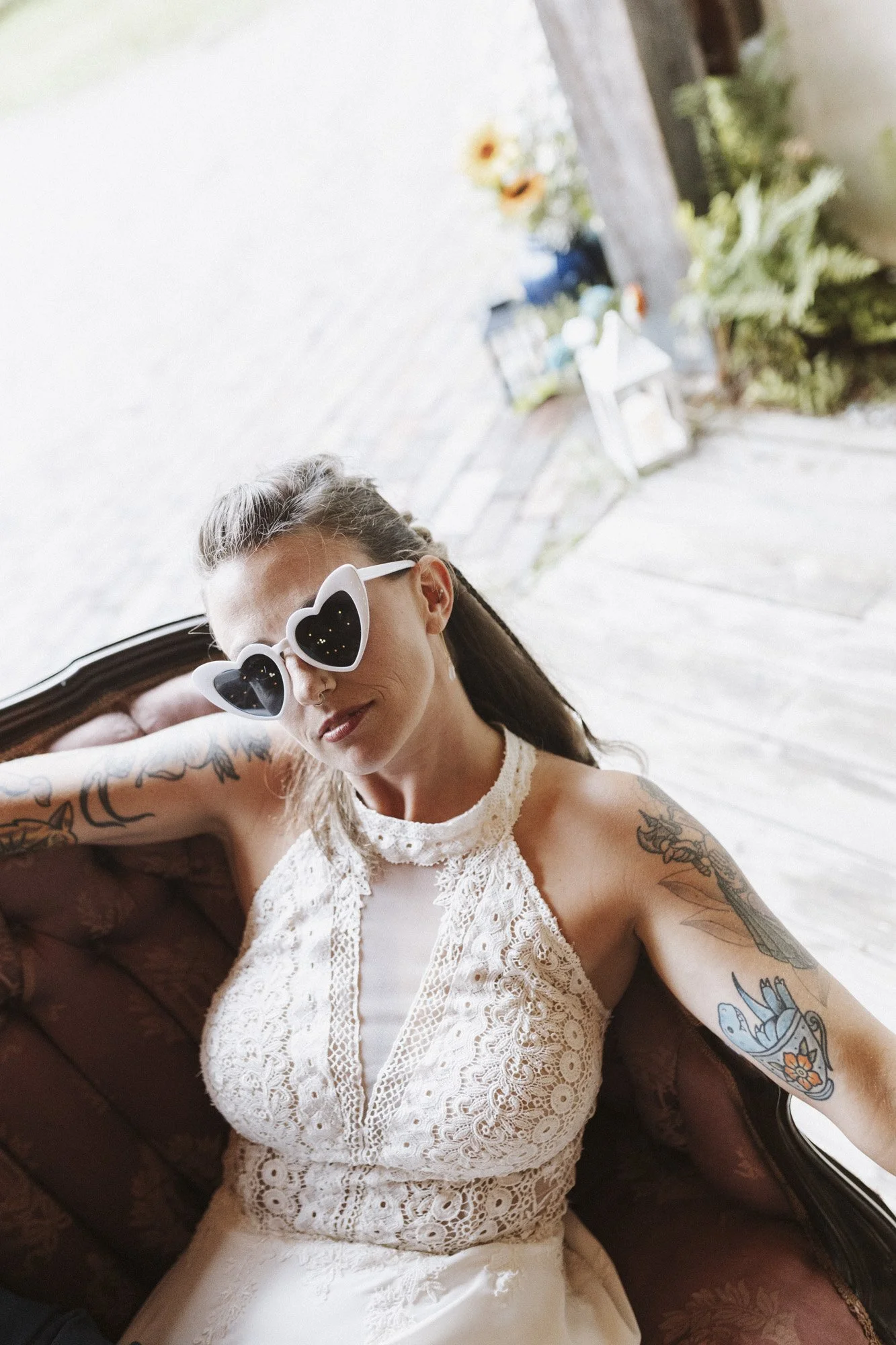 A woman with tattoos on her arms, wearing heart-shaped sunglasses and a white lace dress, sitting on a vintage sofa indoors with a window behind her and an outdoor garden visible.