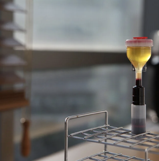 A laboratory setup with a transparent container filled with yellow liquid connected to a black tube, placed on a wire shelf, with a blurred background.