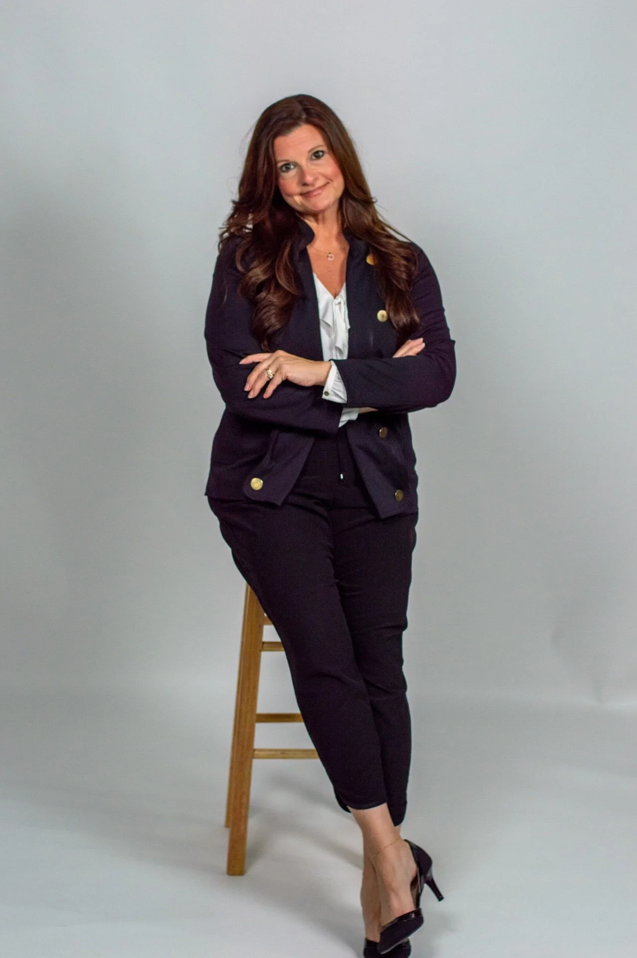 A woman with long, wavy brown hair dressed in a navy blazer, white blouse, and black pants, sitting on a wooden stool against a plain gray background with her arms crossed, smiling at the camera.